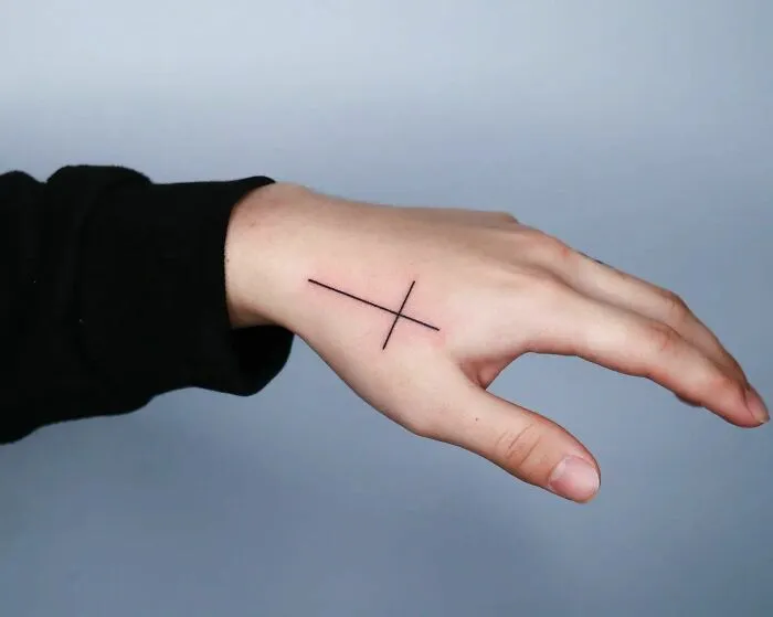 Minimalist small hand tattoo with a simple cross design on the back of a hand.