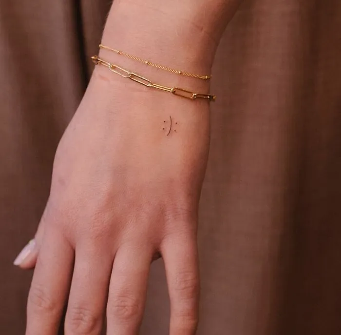 Small hand tattoo of a minimalist smiley face on wrist, alongside gold chain bracelets.