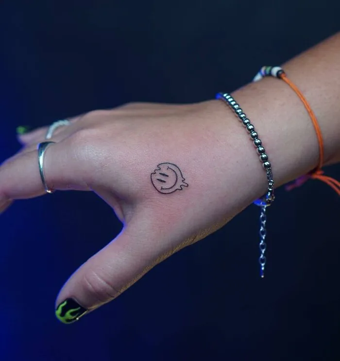Small hand tattoo of a smiley face on the back of a hand with bracelets and nail art visible.