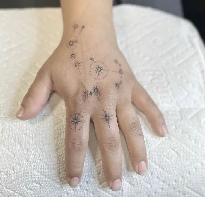Small hand tattoo with constellation design on a person's hand seen on a textured white background.