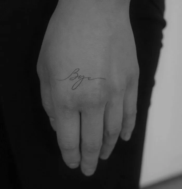 Small hand tattoo with the word "Bye" in elegant script on the side of a finger.