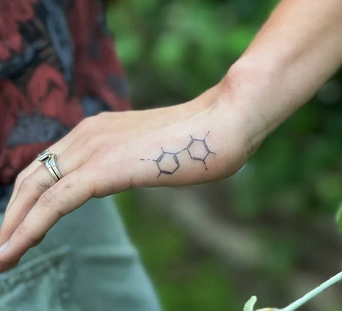 Small hand tattoo of a minimalist geometric design on the wrist, set against a blurred natural background.