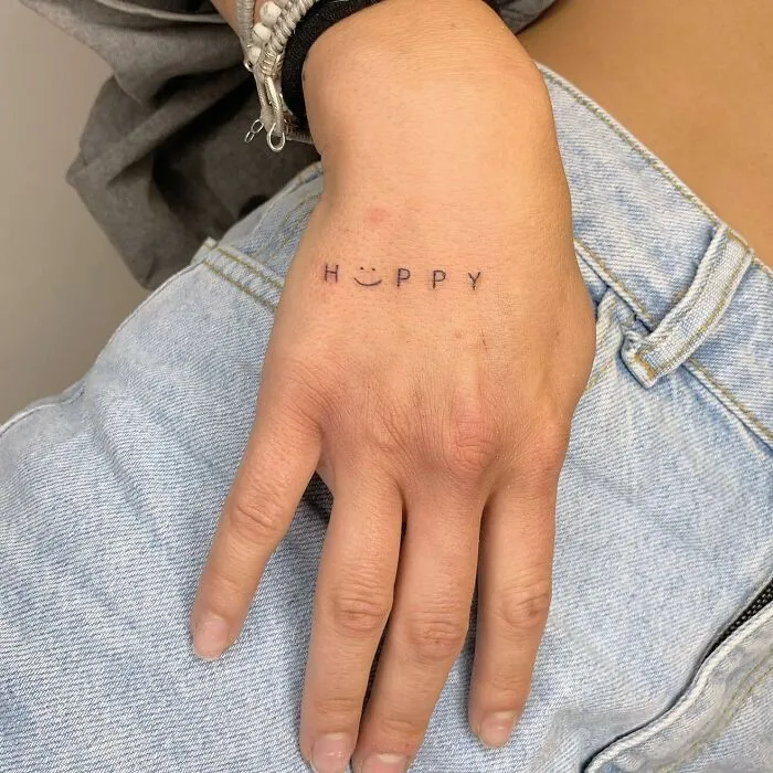Small hand tattoo with the word "HAPPY" and a smiley face on a person's hand wearing jeans.