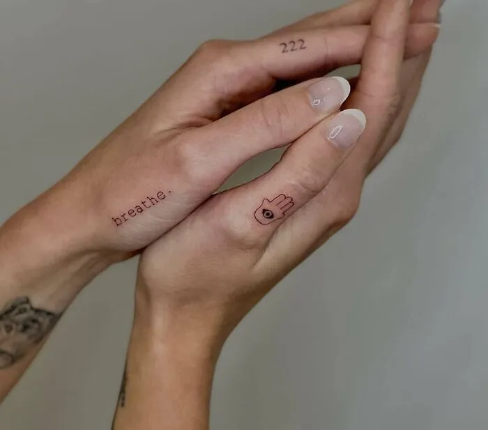 Small hand tattoos featuring the words "breathe" and number "222," with a minimalist eye design on fingers.