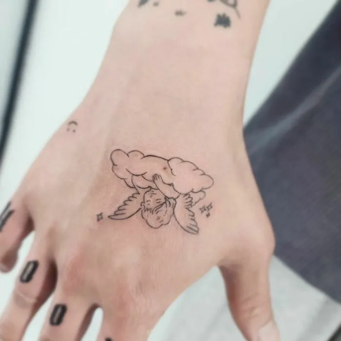 Small hand tattoo featuring an angel nestled in a cloud on the back of the hand.