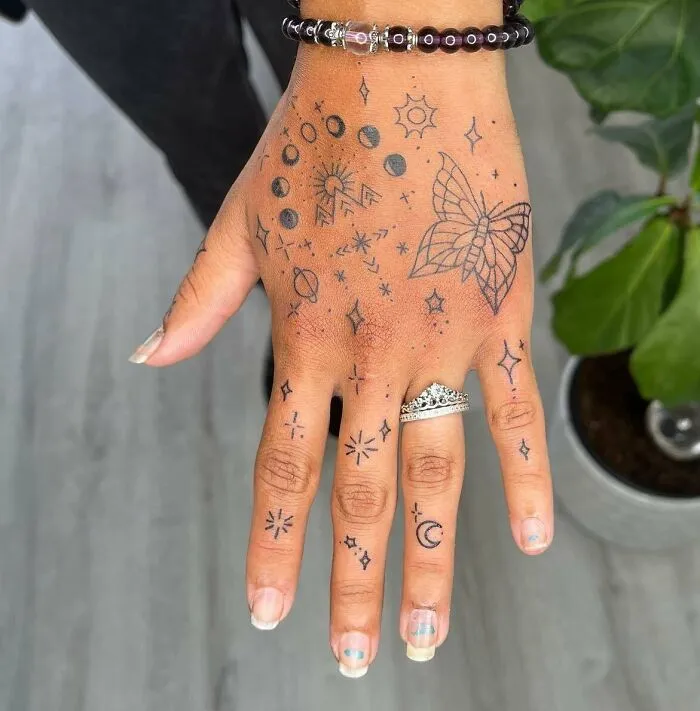 Hand adorned with small tattoos including stars, moon, and a butterfly, showcasing intricate designs on fingers and back of the hand.
