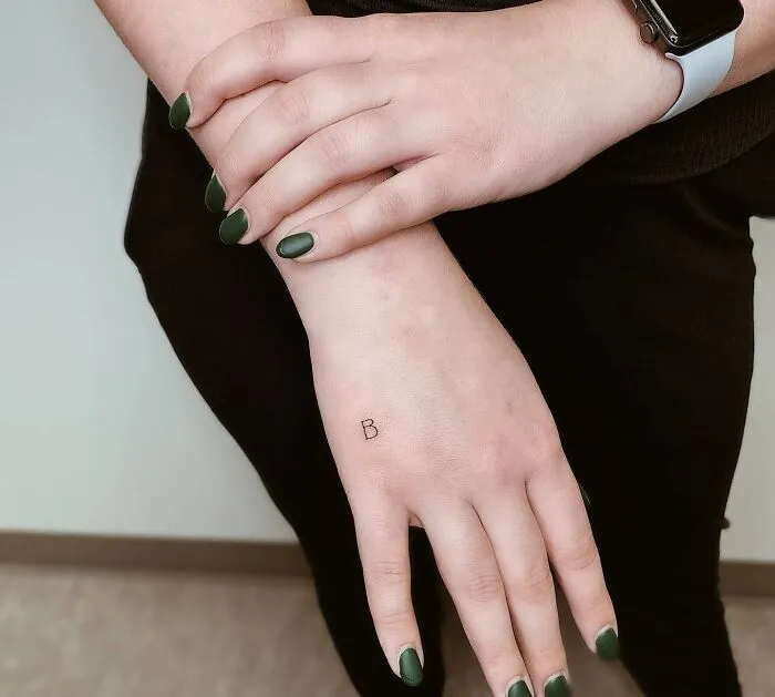 Small hand tattoo of letter "B" on wrist, with green nail polish and a black outfit.