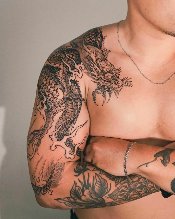 Black dragon tattoo on a man's arm and shoulder, showcasing intricate designs and bold lines.