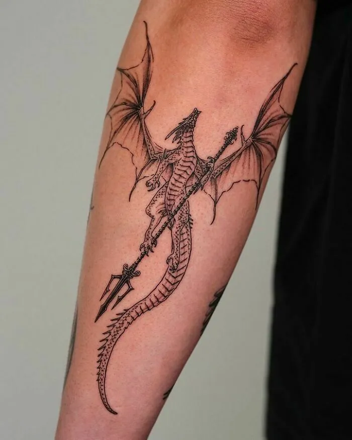 Dragon tattoo design with spear on forearm, showcasing intricate wing and tail details.