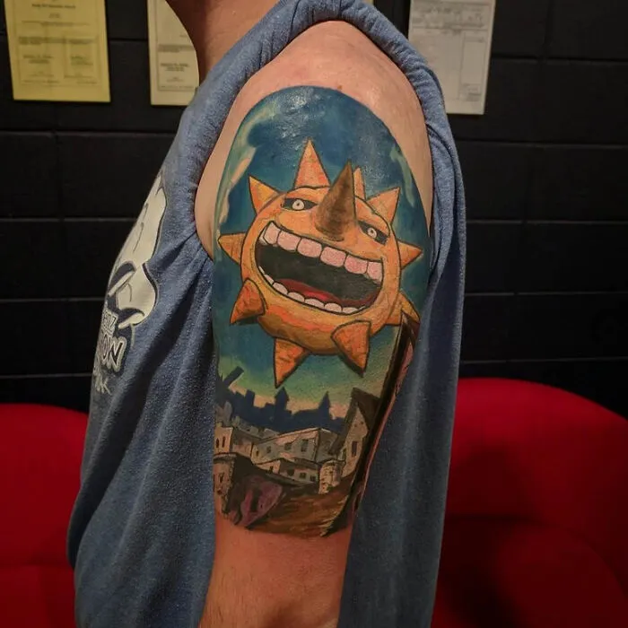 Anime tattoo on upper arm featuring a grinning sun with a background of a townscape and blue sky.