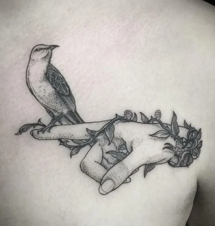 Collarbone tattoo of a detailed bird perched on a hand, entwined with floral vines.