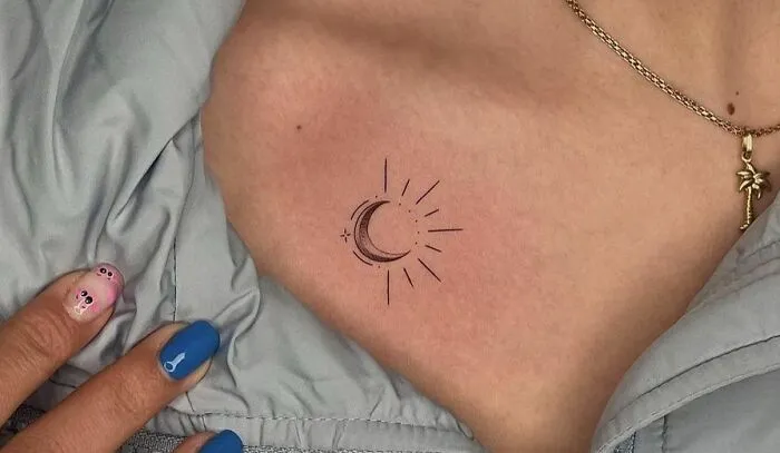 Minimalist crescent moon collarbone tattoo adorned with delicate lines, complemented by blue and pink nail art.