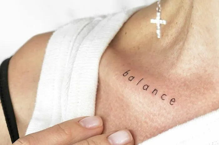 Collarbone tattoo with the word "balance" in black ink on a person's skin, next to a cross earring.