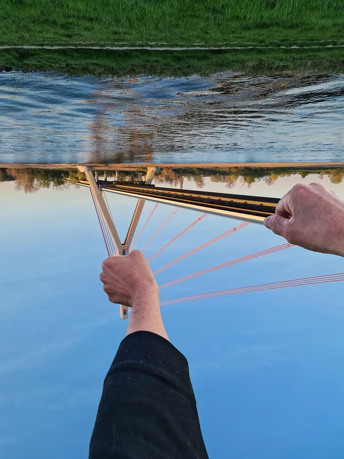 Playing with perspective: Two hands appearing to hold the cables of an inverted suspension bridge over a river, changing what we see.