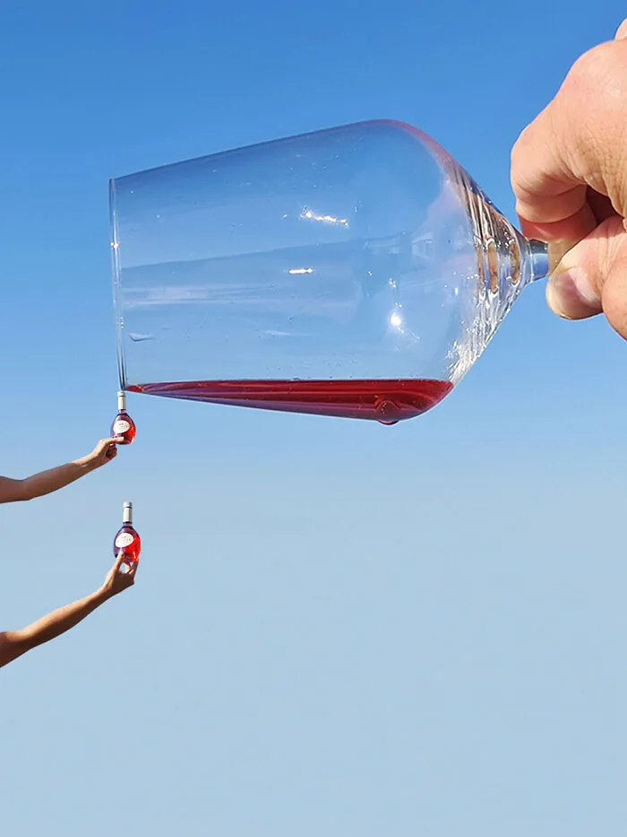 Playing with perspective: A giant wine glass held by a hand, pouring red wine into tiny bottles held by other hands, changing what we see.