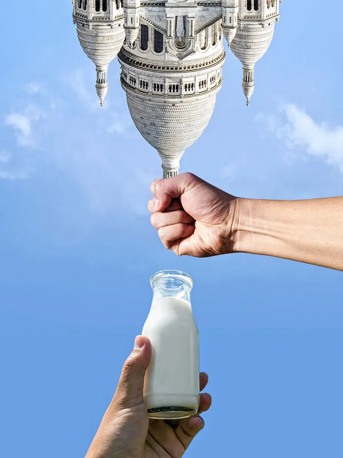 An upside-down building appears to be held by a fist, with another hand holding a milk bottle below, playing with perspective.