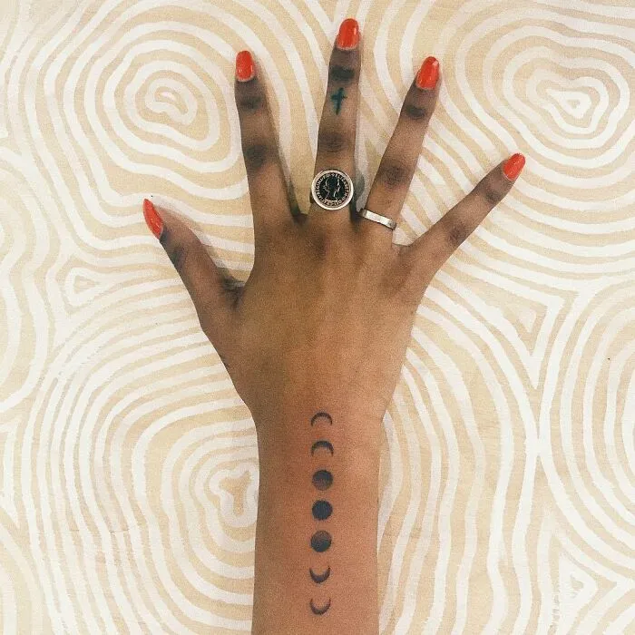 Hand with red nails showing moon phase wrist tattoo design.