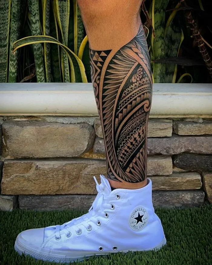 Leg with tribal tattoo design, wearing white Converse sneakers, standing on grass with stone wall background.