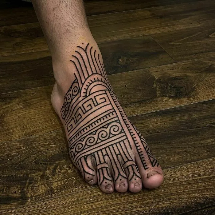 A foot showcasing an intricate tribal tattoo design with geometric patterns on a wooden floor.