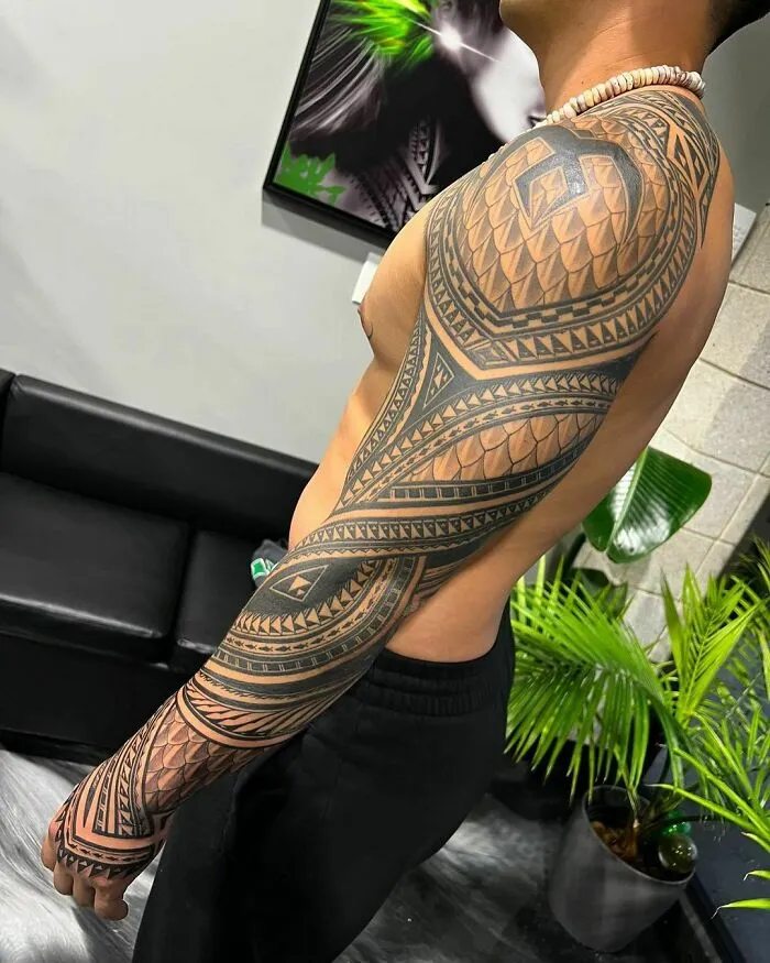 Man with a tribal tattoo sleeve on bare arm in a studio setting.