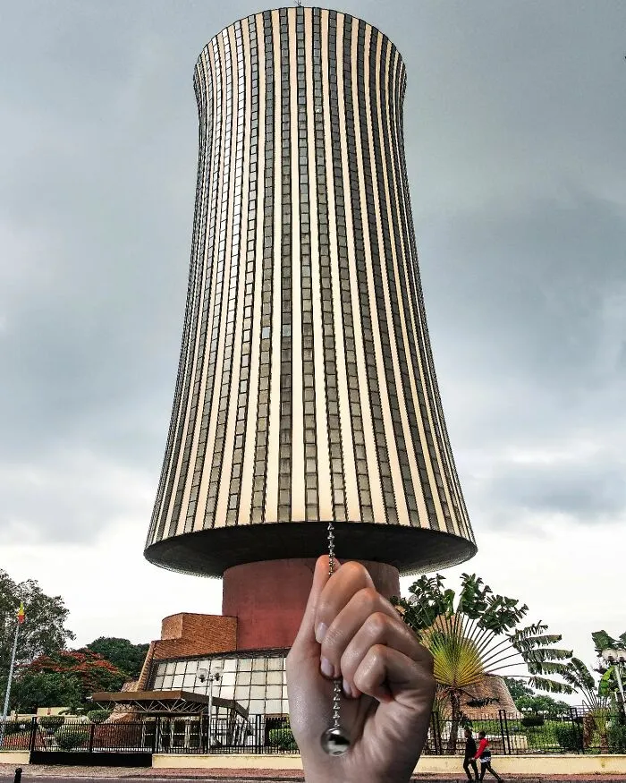 A hand holds a pull chain against a tall, cylindrical building, making the building look like a light fixture to change perspective.