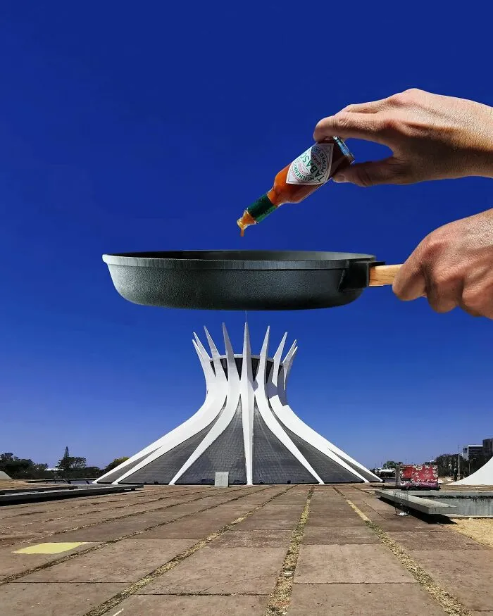 A hand holds a frying pan over a distinctive white building with a hand squeezing hot sauce into it, creating a playful change in perspective.