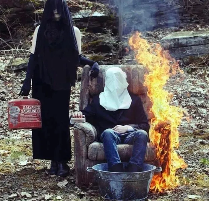 Cursed meme of a woman in black veil holding gas can, standing next to a burning seated figure with covered face.