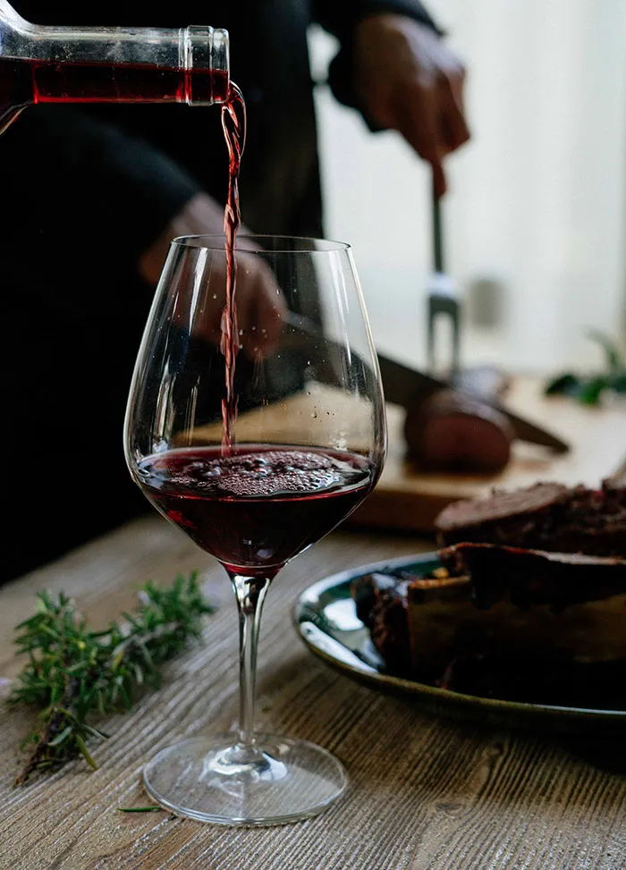 Red wine pouring into a glass, with a person carving meat in the background, representing food history and dining.