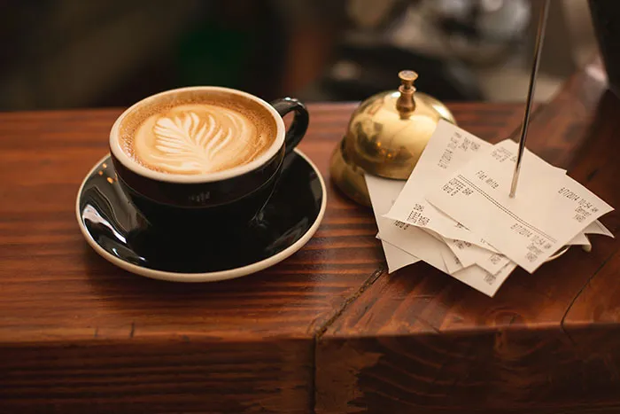 A cup of coffee with latte art next to a bell and stack of receipts, showcasing food history.