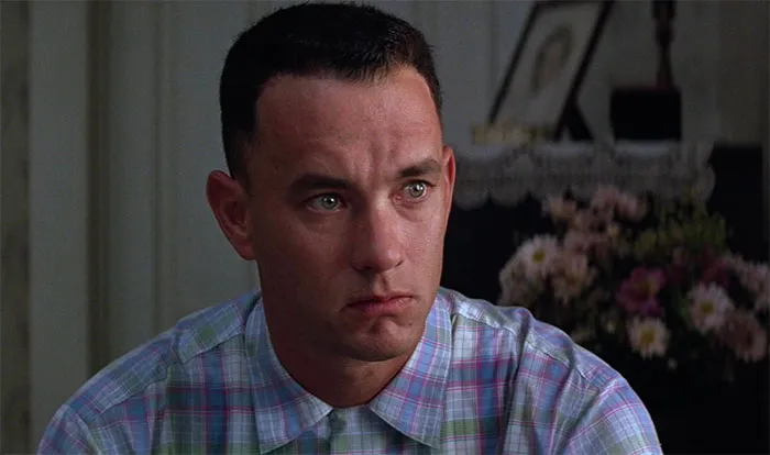 Forrest Gump wearing a plaid shirt, looking thoughtful in a dimly lit room with floral decorations behind him.