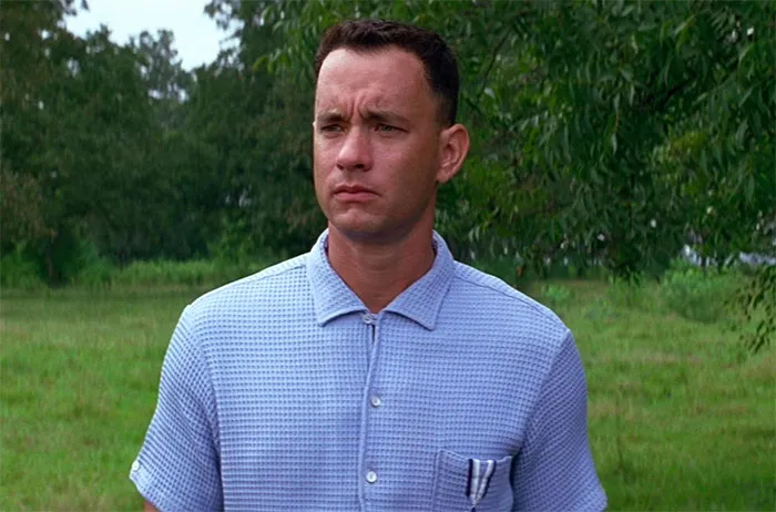 Forrest Gump wearing a blue shirt standing outdoors with trees and grass in the background for Forrest Gump quotes content.