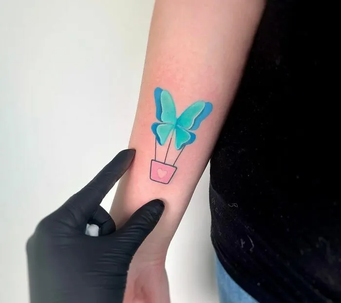 Colorful butterfly tattoo with a teal design and heart on forearm, being showcased by a person wearing a glove.