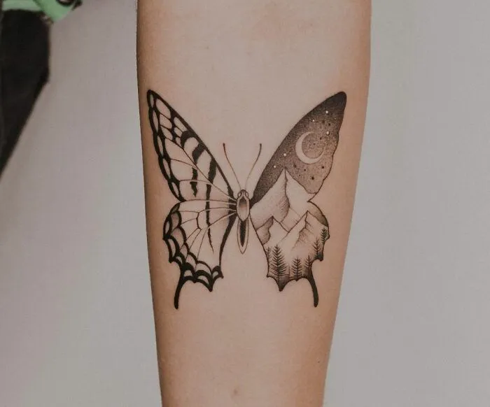Butterfly tattoo with intricate black and white design featuring a mountain and forest scene on one wing.