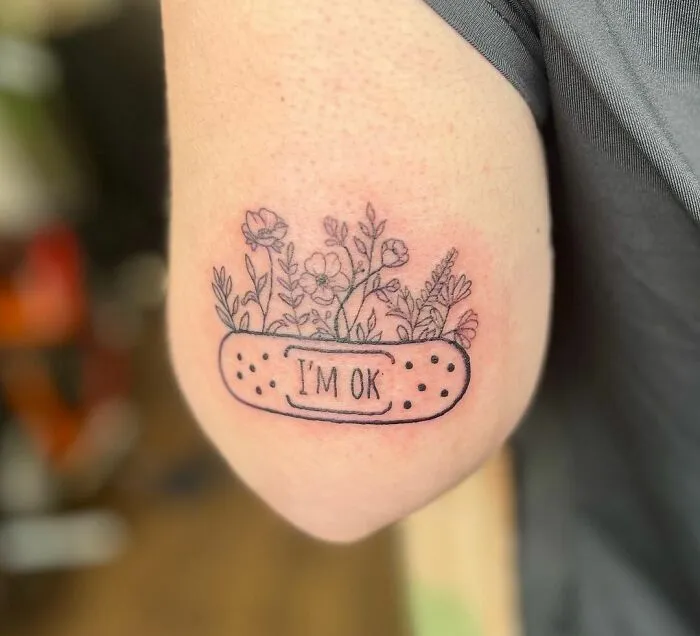 Tattoo of a bandage with flowers and the words "I'm OK," symbolizing mental health and healing on a person's arm.