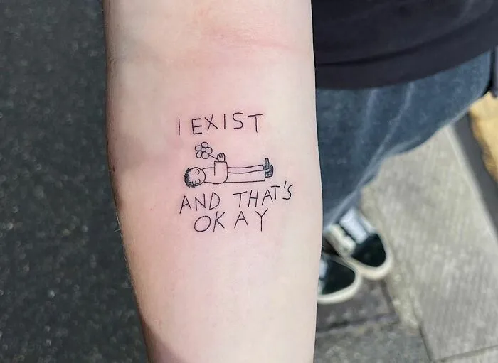 Mental health tattoo on forearm with text "I exist and that's okay," featuring a small figure holding a flower.