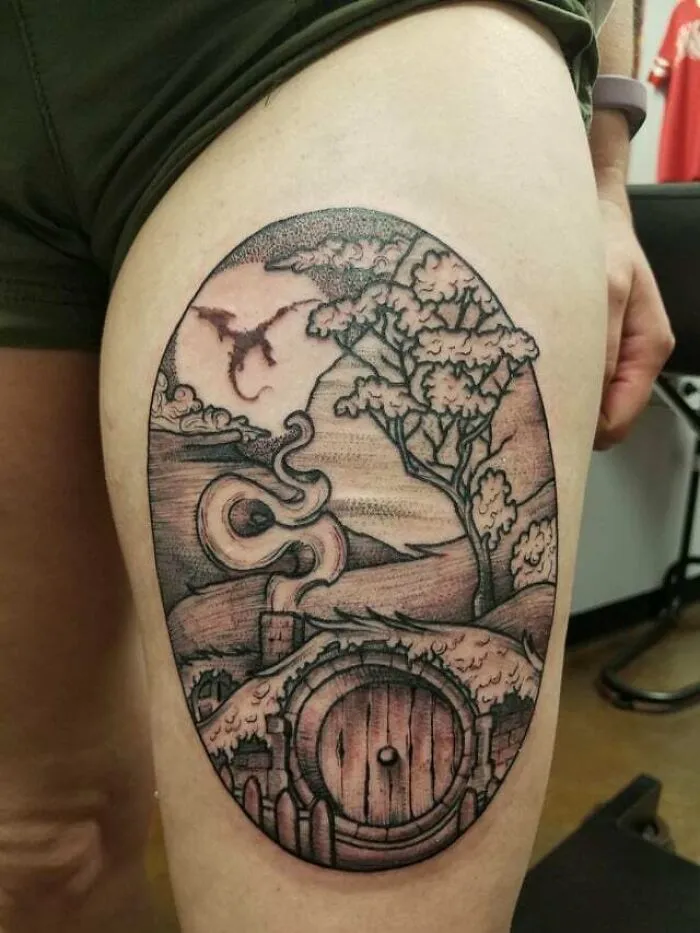 Lord of the Rings tattoo featuring a Hobbiton scene with a dragon, detailed on a person's thigh.