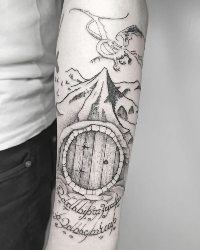 Lord of the Rings tattoo featuring a dragon, mountain, and round door with Elvish script on an arm.