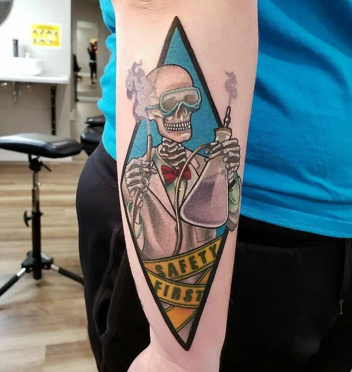 Science tattoo of a skeleton scientist with goggles and flasks, emphasizing safety, on a person's forearm.