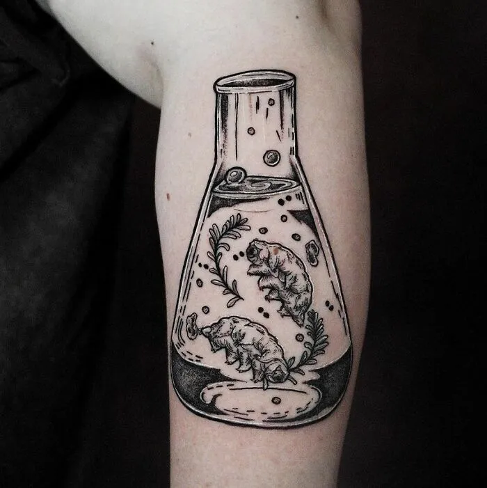 Science tattoo of a beaker with tardigrades and foliage, inked on forearm.