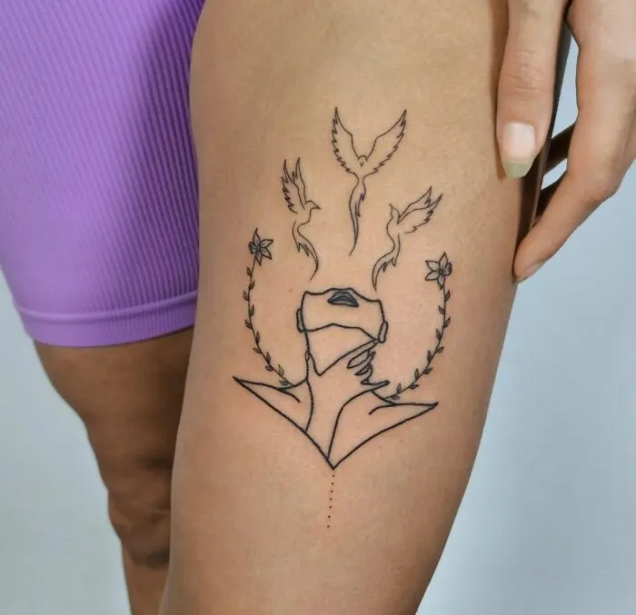 Minimalist mental health tattoo with a face, birds, and floral elements on a thigh in a healing motif.
