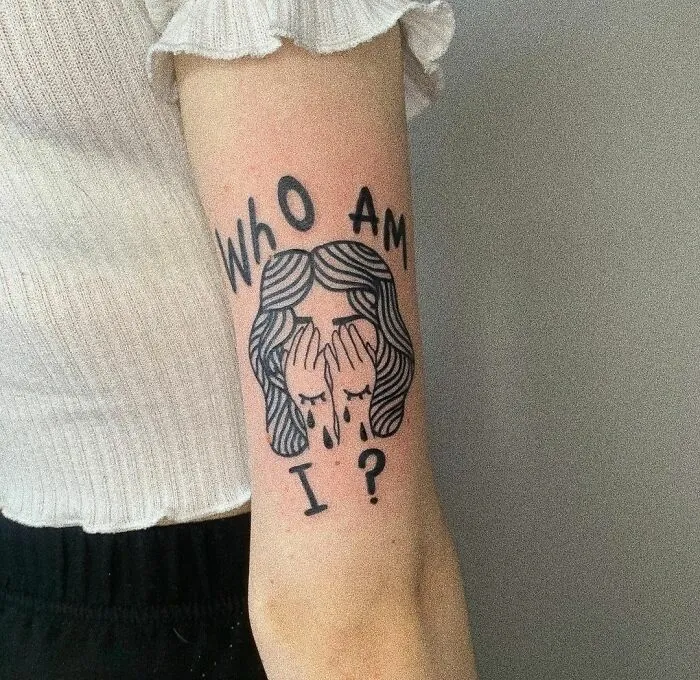 Tattoo on arm asking "Who Am I?" with a face covering hands, symbolizing mental health exploration.