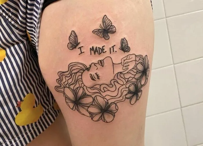 Mental health tattoo with a woman's face, surrounded by butterflies and flowers, with the text "I made it" on the thigh.