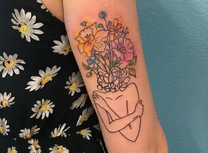 Arm tattoo featuring a mental health theme: a figure embracing themselves with colorful flowers as the head.