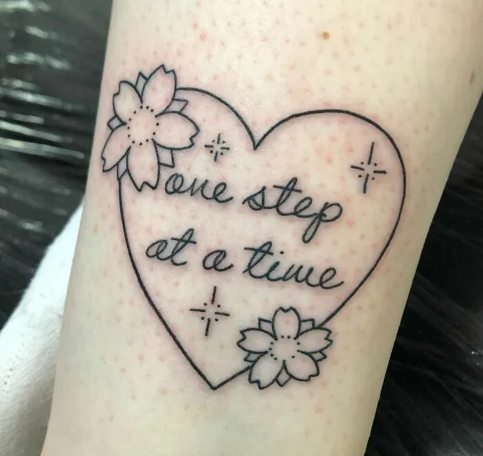 Heart-shaped mental health tattoo with "one step at a time" text and floral accents.