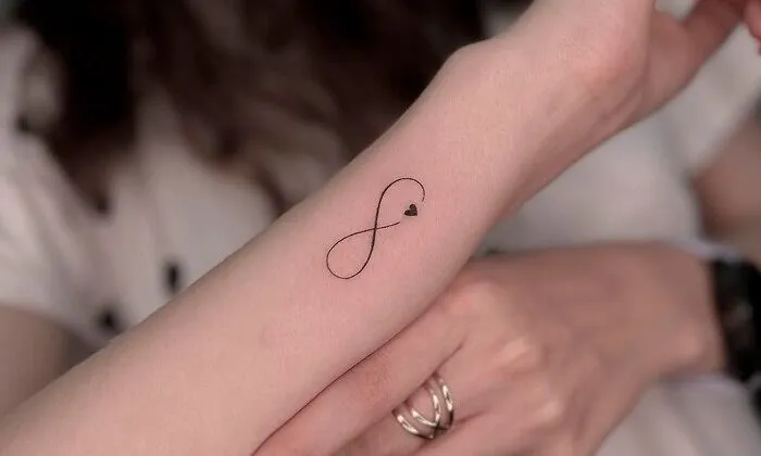 Minimalist infinity tattoo with a small heart on a forearm, symbolizing eternal creativity.