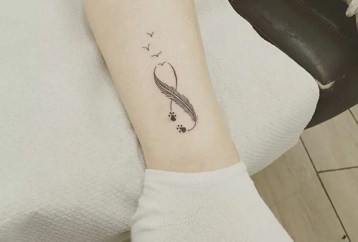 Infinity tattoo with feather and bird elements on arm, showcasing eternal creativity.