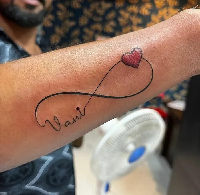 An infinity tattoo with a heart and name design on an arm, symbolizing eternal creativity.