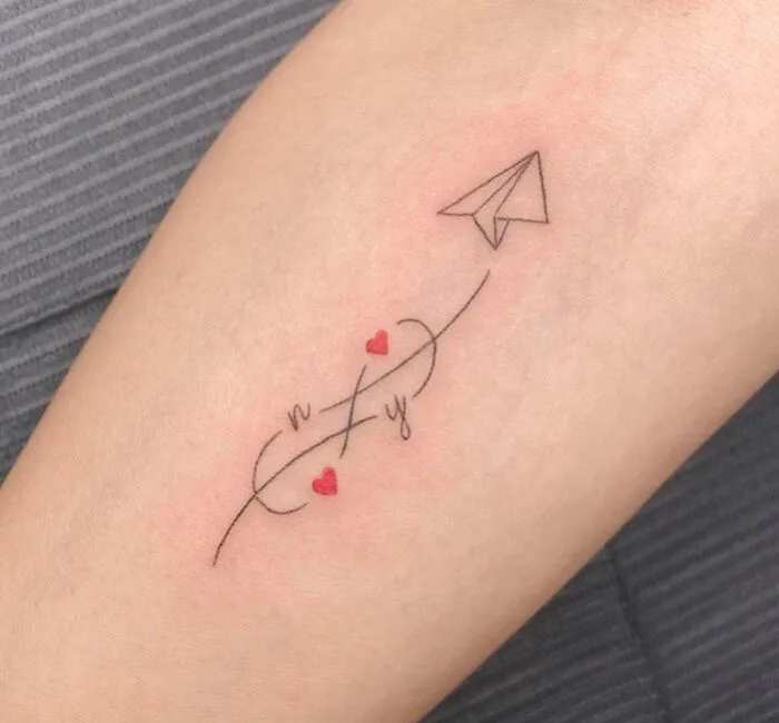 Infinity tattoo with paper airplane and red hearts on arm, symbolizing eternal creativity.