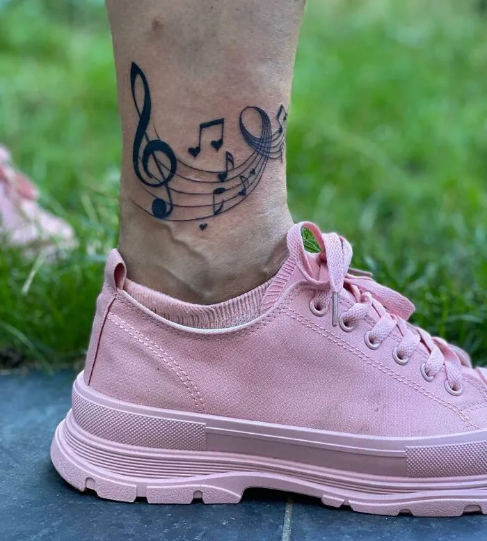Music tattoo on ankle with musical notes and symbols, worn with pink sneakers, displayed outdoors on grass.