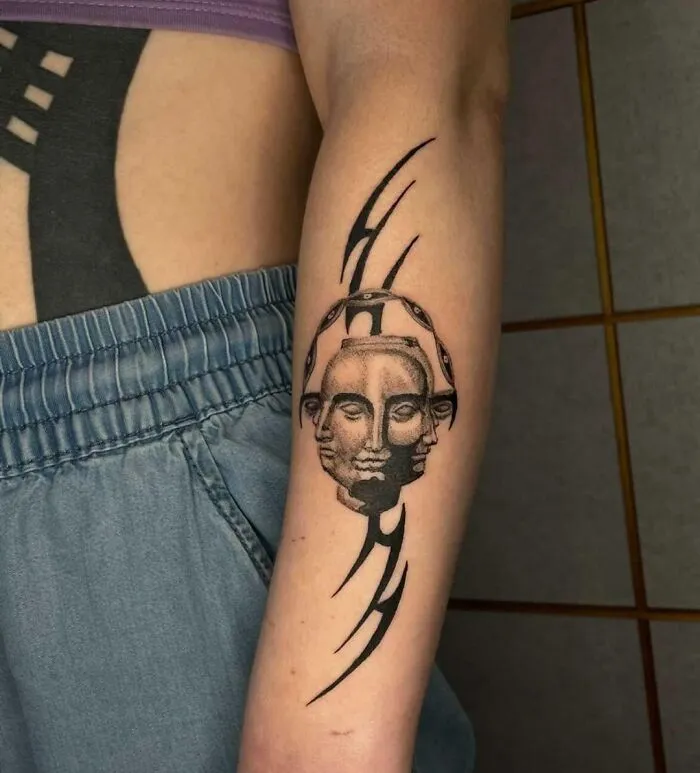 Arm with tribal tattoo featuring a double-faced figure and curved lines, worn with blue shorts.